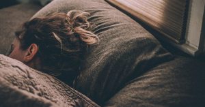 Woman asleep under gray blankets as morning light comes through the blinds, illustrating feeling tired all day wired at night